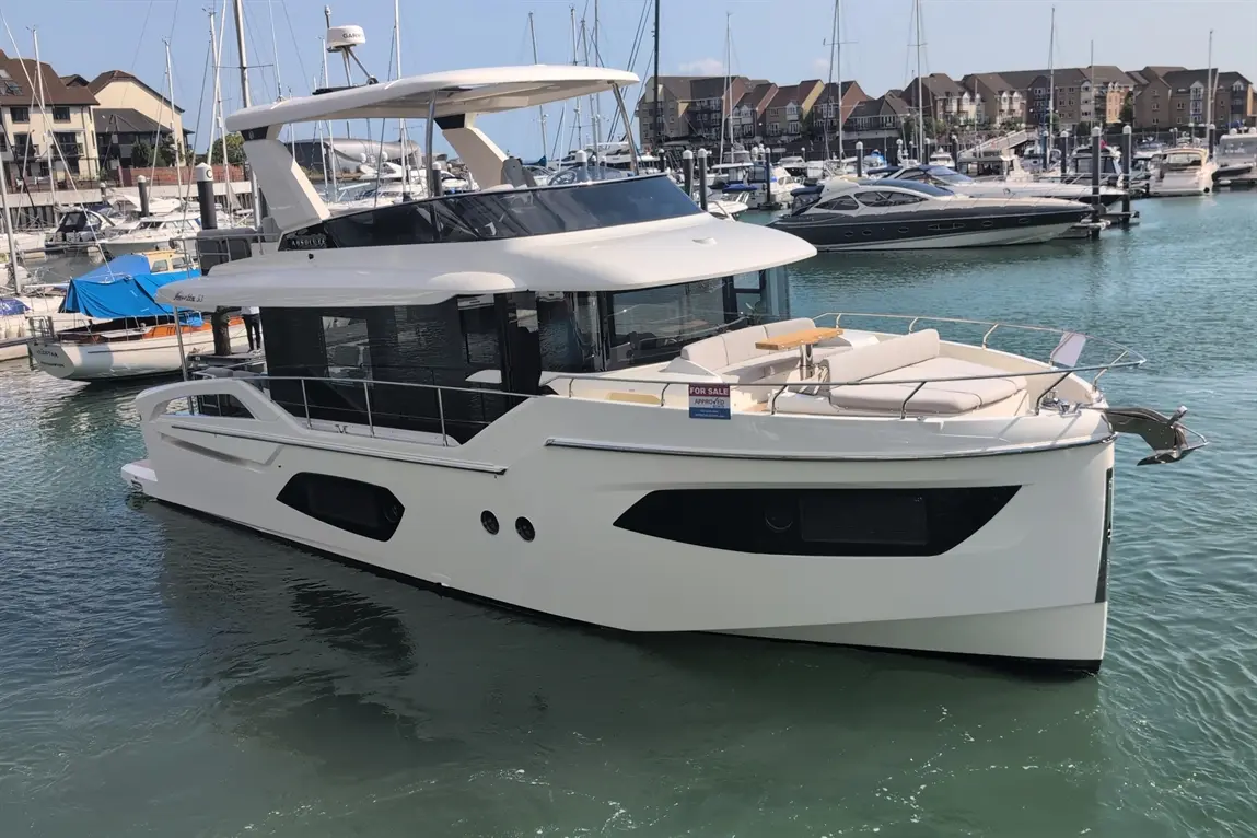 A modern white yacht with a spacious deck is docked at a marina, featuring a sign reading "FOR SALE" on the side.